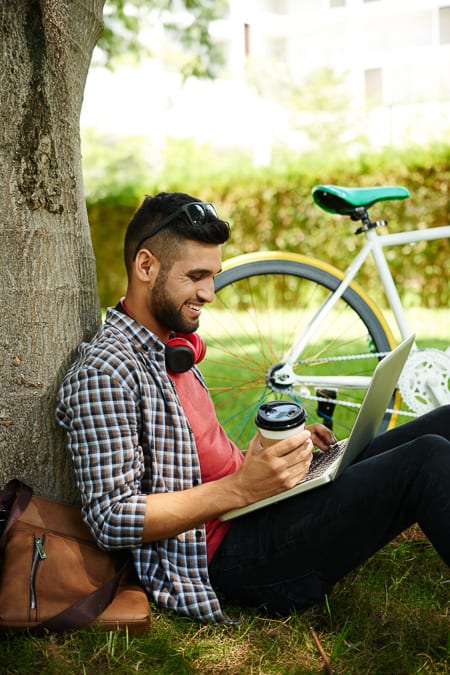 man with laptop and coffee leaning against a tree with a bike next to him