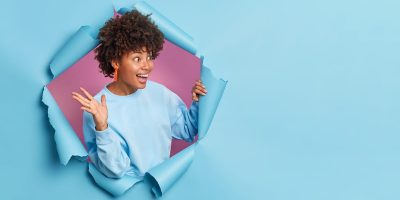 Overjoyed African American woman raises palm and looks gladfully aside as meets someone wears casual jumper poses through paper background in hole expresses positive emotions.